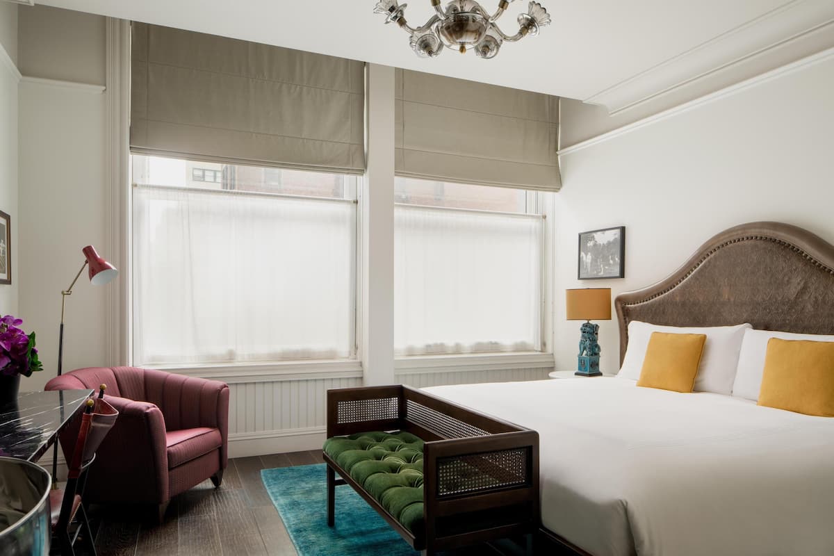 Quarto de hotel com cama, uma cadeira, uma mesinha, luminária e janela com persianas.