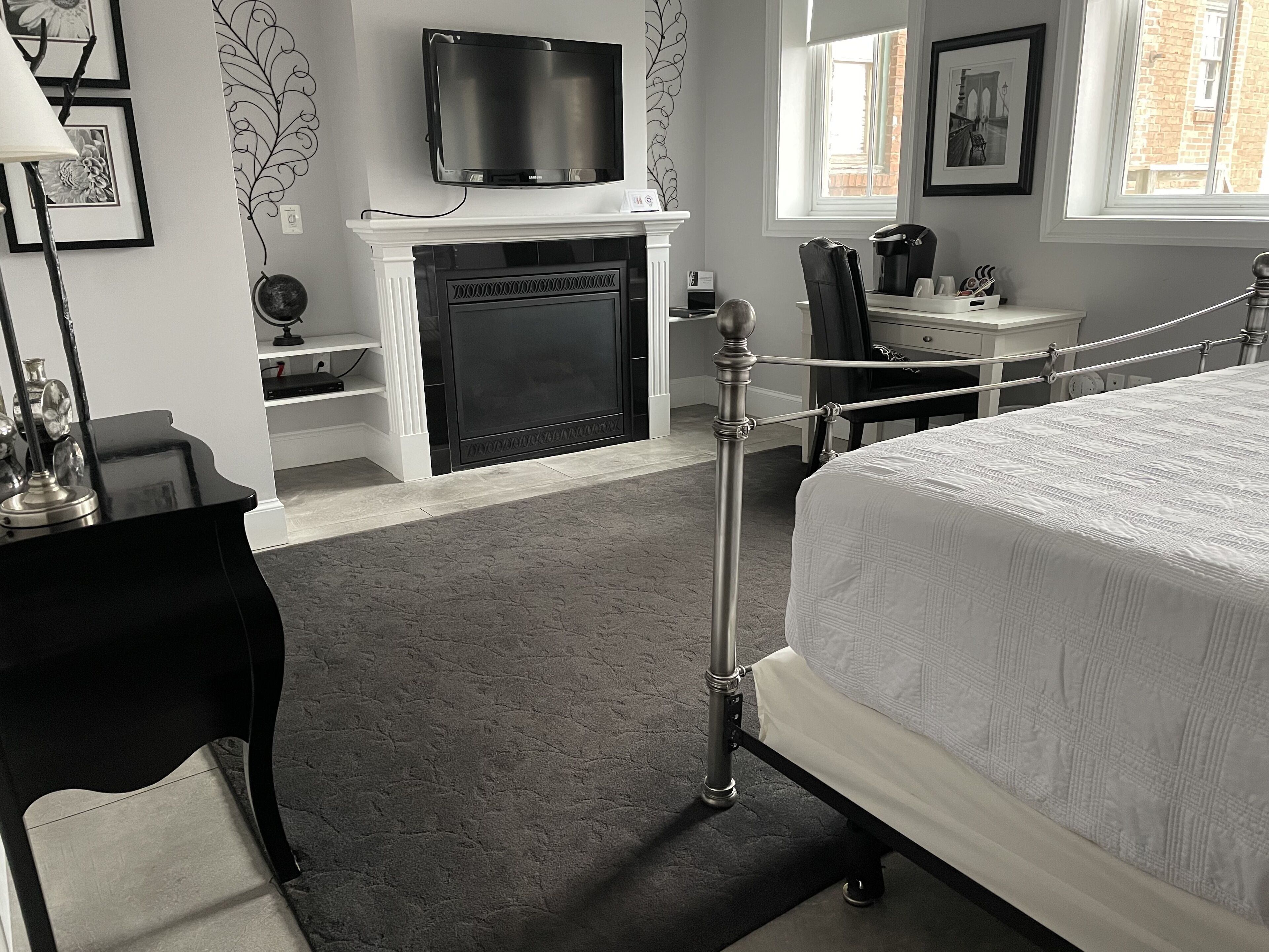 A bedroom with a bed, a television mounted above a fireplace, a desk with a chair, and a window with a view of a brick building.