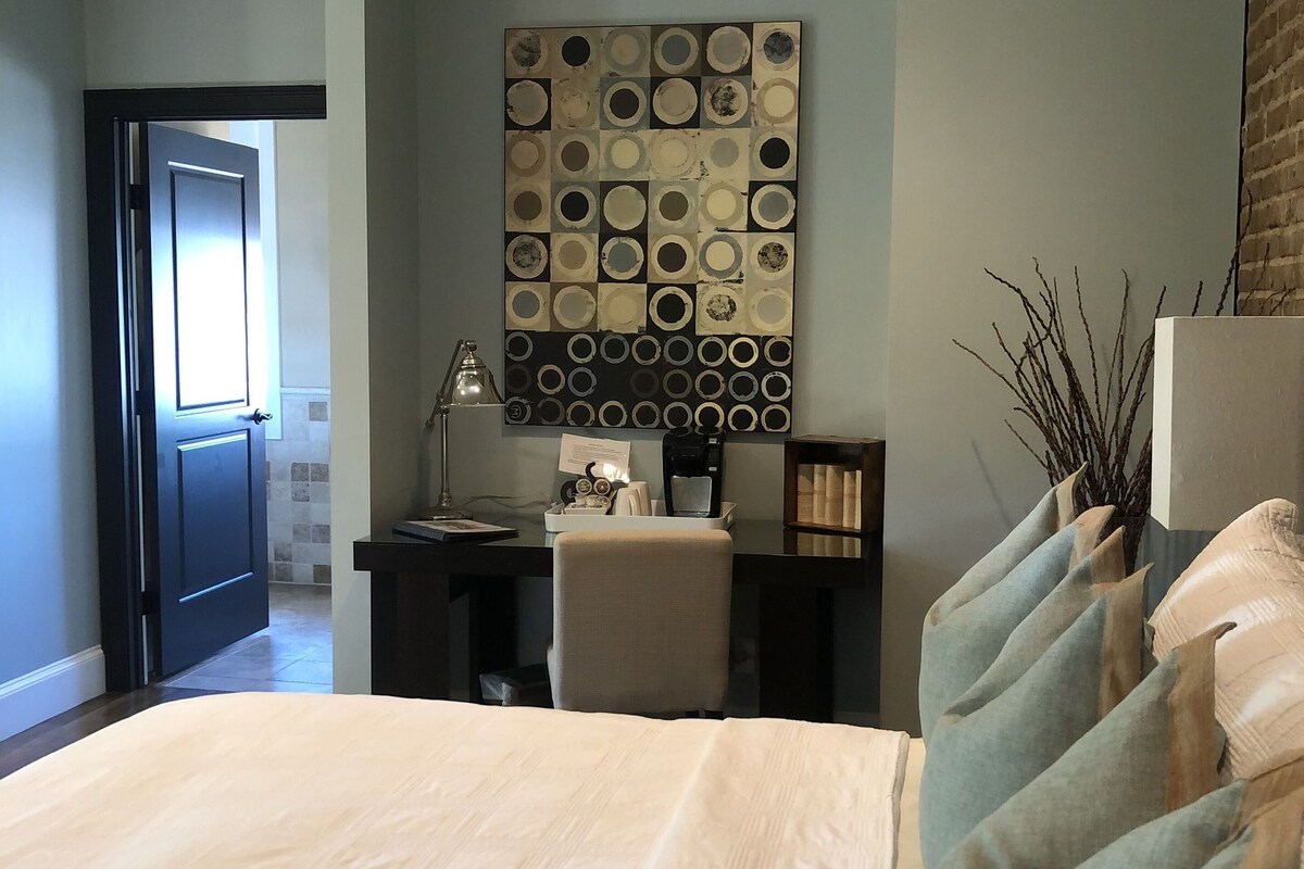 A bedroom with a large bed, a desk with a chair, a coffee maker, and a decorative wall piece.