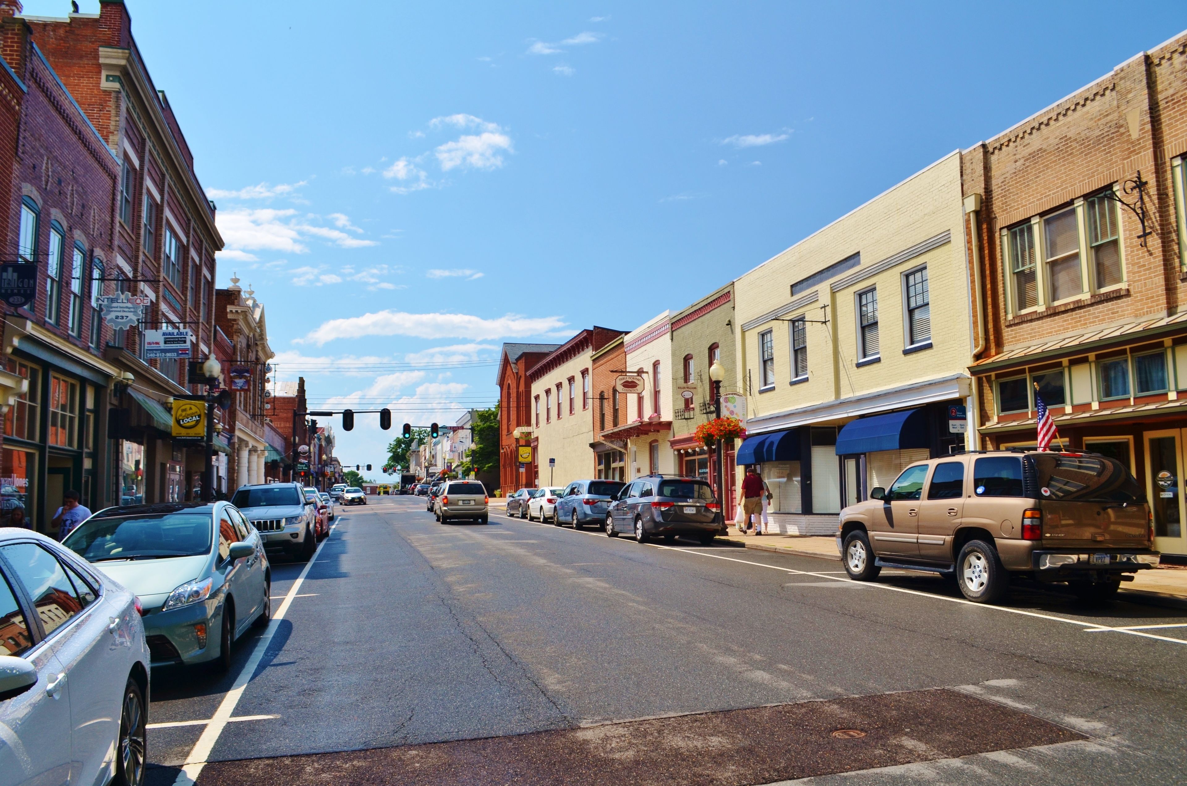 A street lined with multi-story buildings, parked cars, and a clear blue sky.