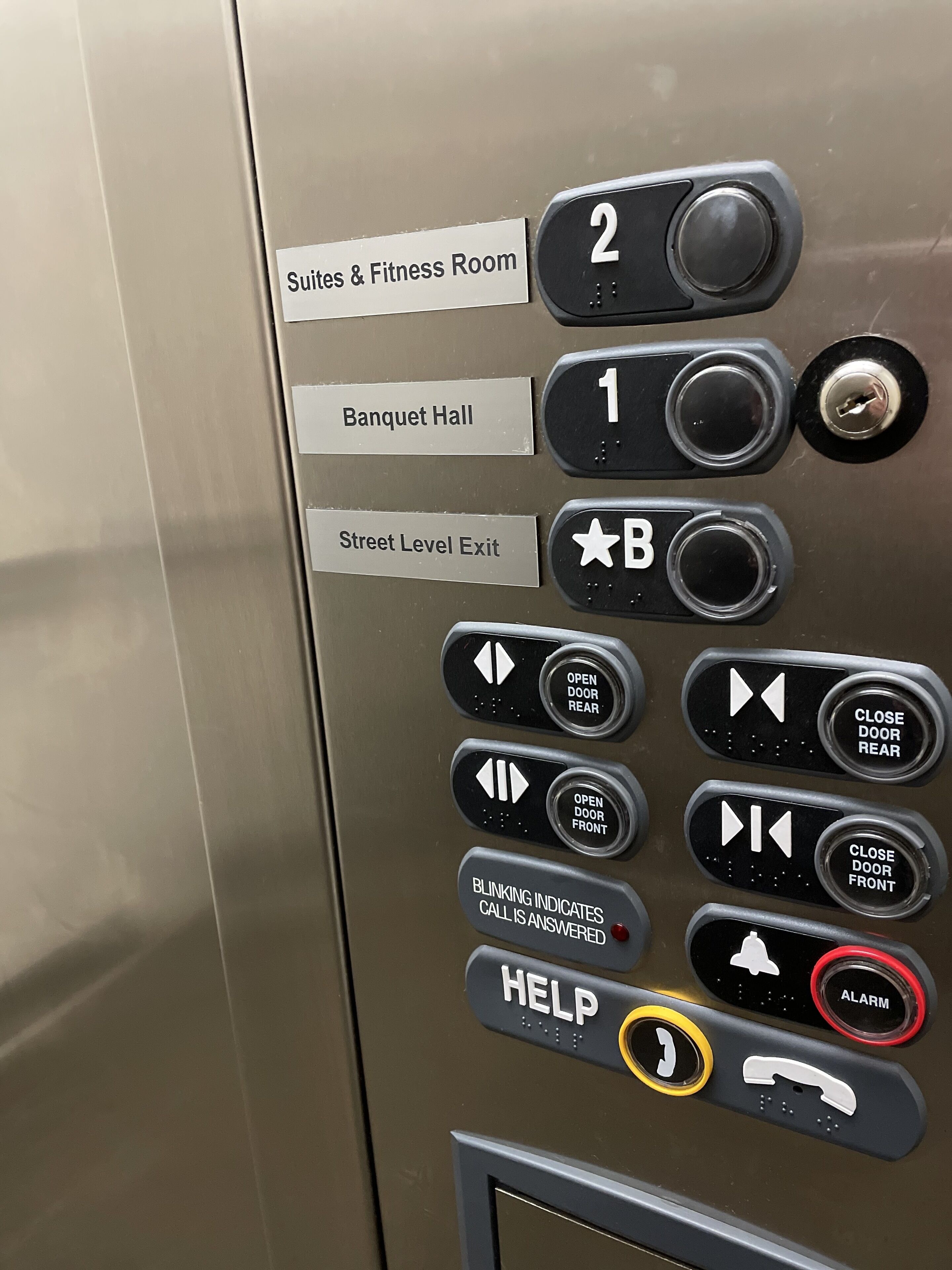 Panel de control de ascensor con botones para 'Suites & Fitness Room', 'Salón de Banquetes' y 'Salida a Nivel de Calle'.
