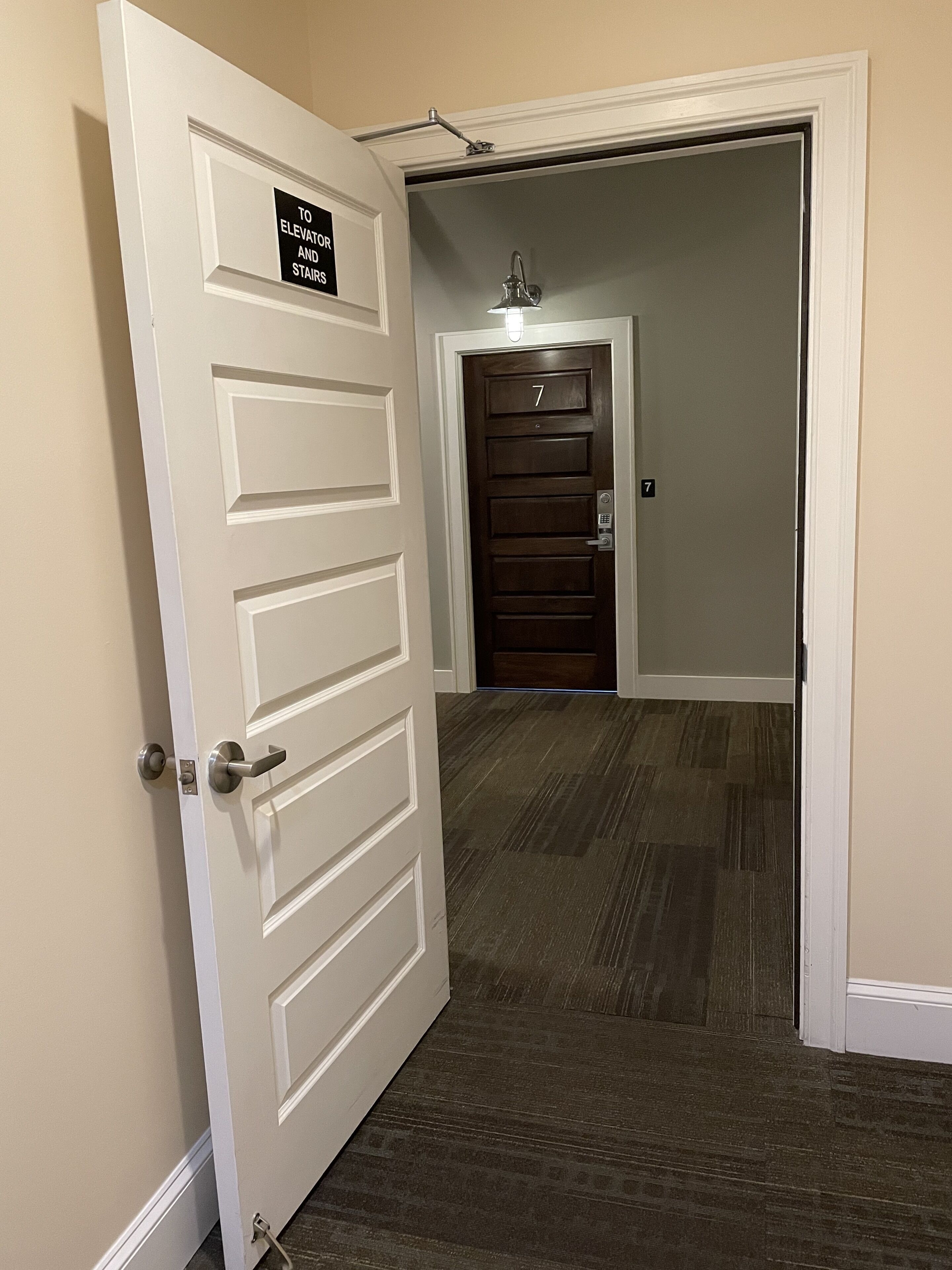 Un couloir d’hôtel avec un sol recouvert de moquette, une porte blanche portant une indication signalant la direction de l’ascenseur et des escaliers, et une porte portant le numéro 7.
