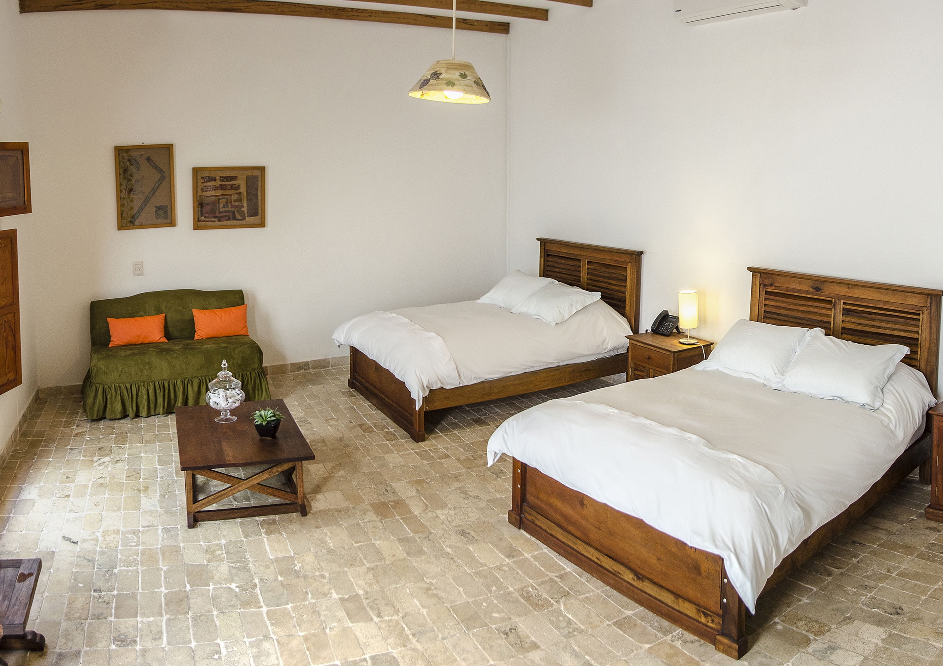A room with two wooden beds, a green sofa with orange cushions, a small table with a glass jar and a plant, and a hanging light fixture.