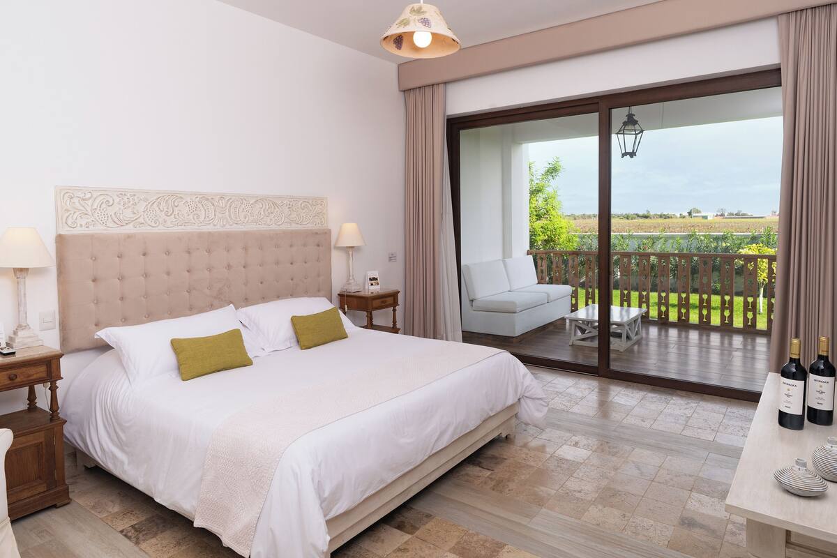 Une chambre avec un grand lit, une table de chevet, une commode, une chaise, un balcon avec vue et une table avec des bouteilles.