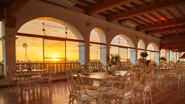 An outdoor dining area with wicker chairs and tables, large arched windows, and a view of the sunset.