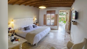 A bedroom with a large bed, a desk with a chair, a TV mounted on the wall, and a view of the garden through the open window.