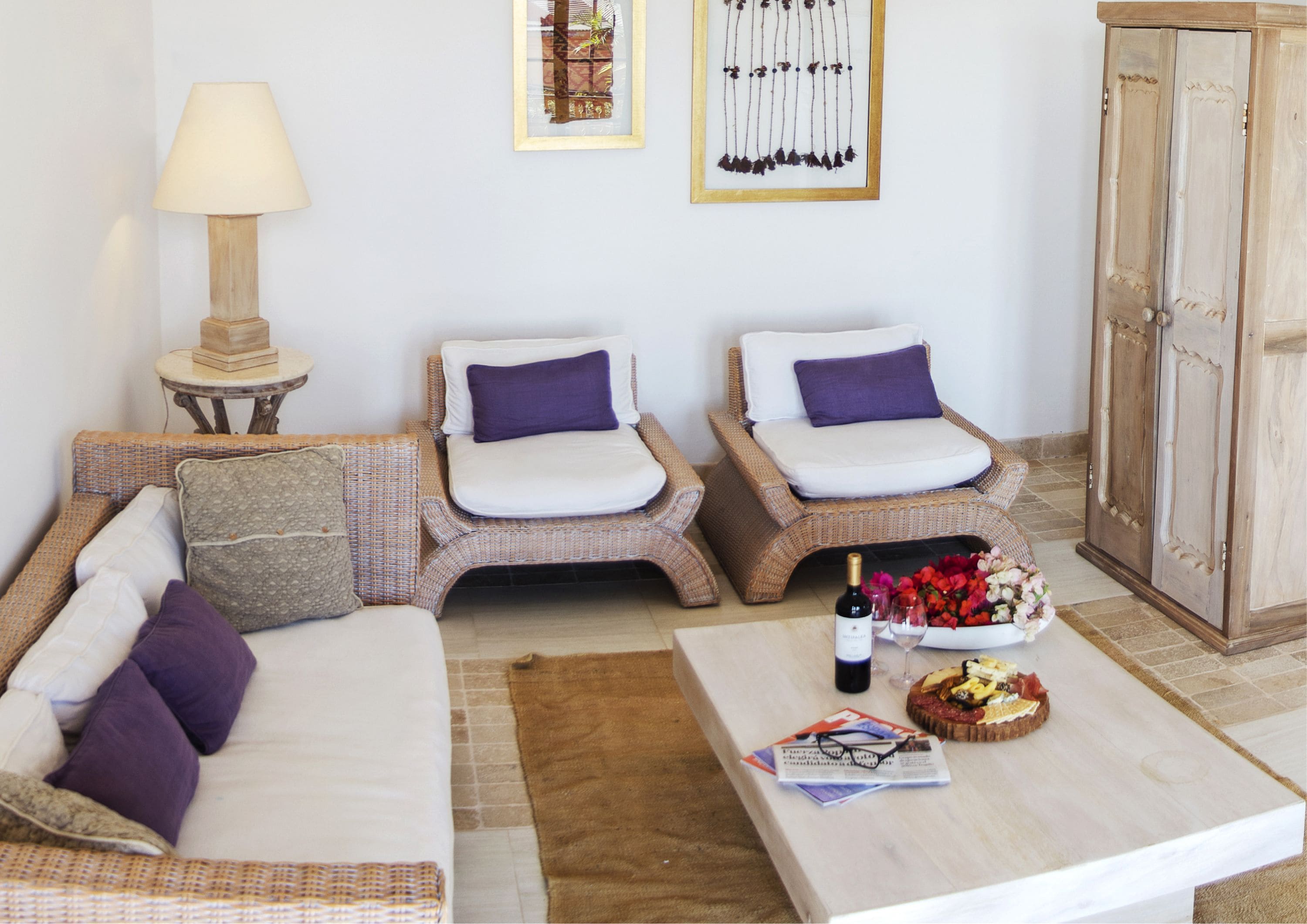 A cozy room with a sofa, two wicker chairs, a small table with a bottle and flowers, and a cabinet.