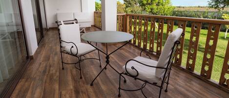A balcony with a round glass table and two white cushioned chairs, wooden flooring, and a view of greenery.