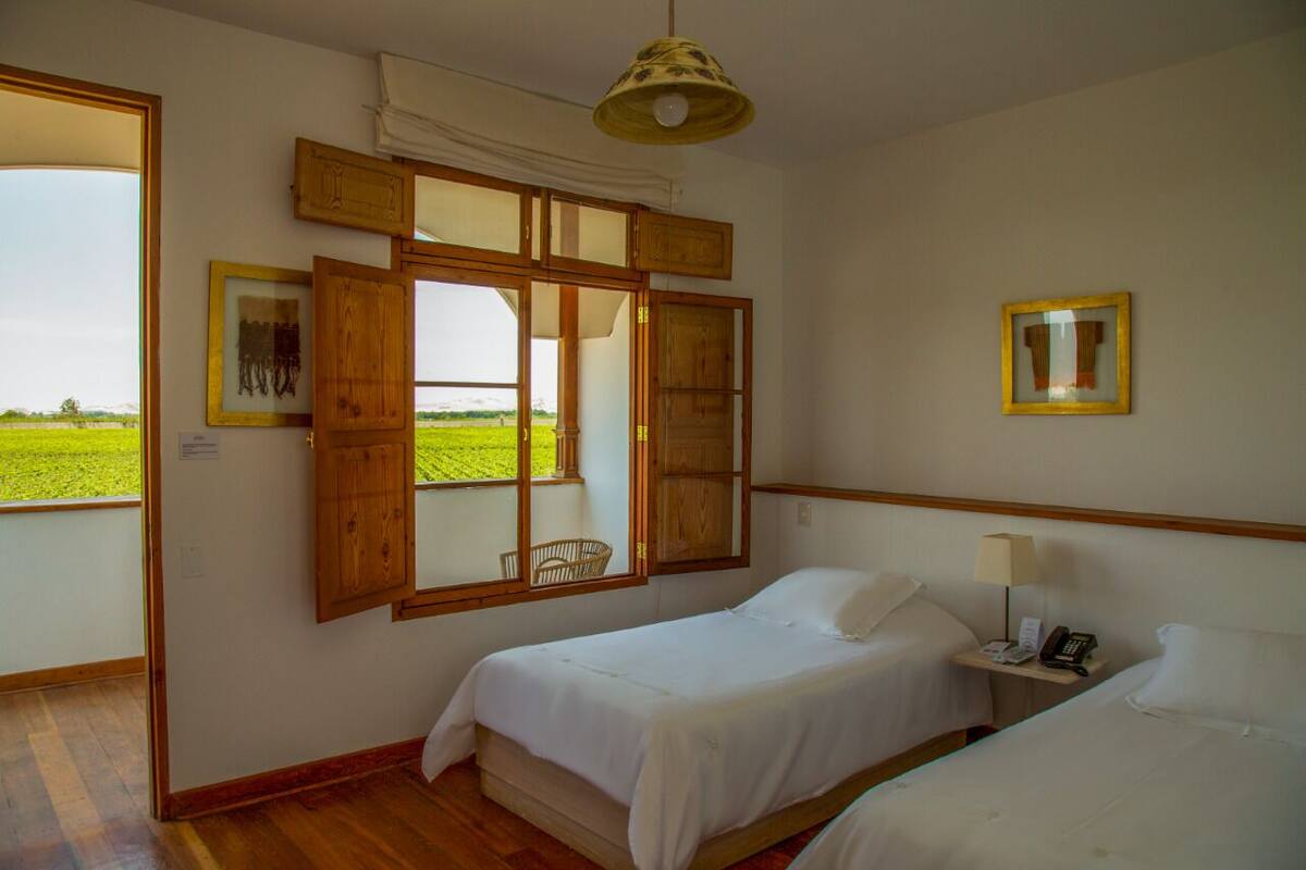 Une chambre avec deux lits, une fenêtre avec des volets en bois, une lampe suspendue et une petite table de chevet avec une lampe et un téléphone.