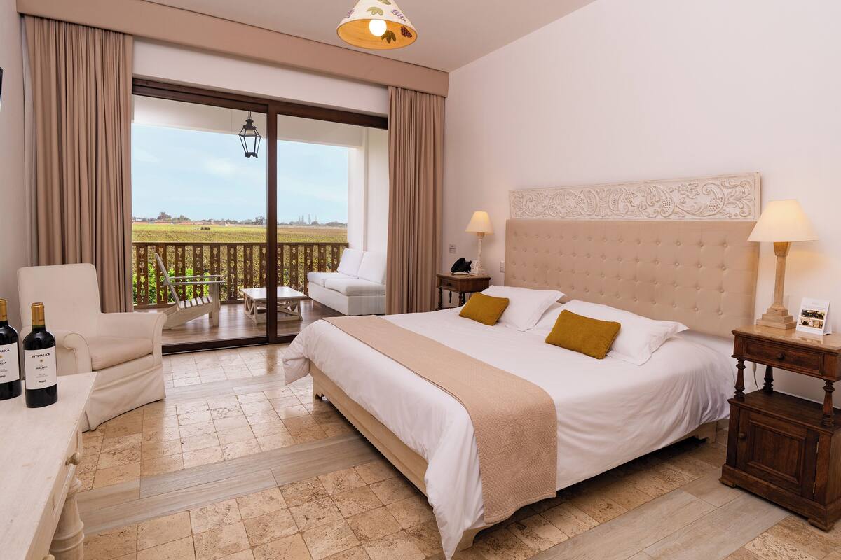 Une chambre d’hôtel avec un lit, une chaise, une table, un balcon avec vue et deux bouteilles de vin.