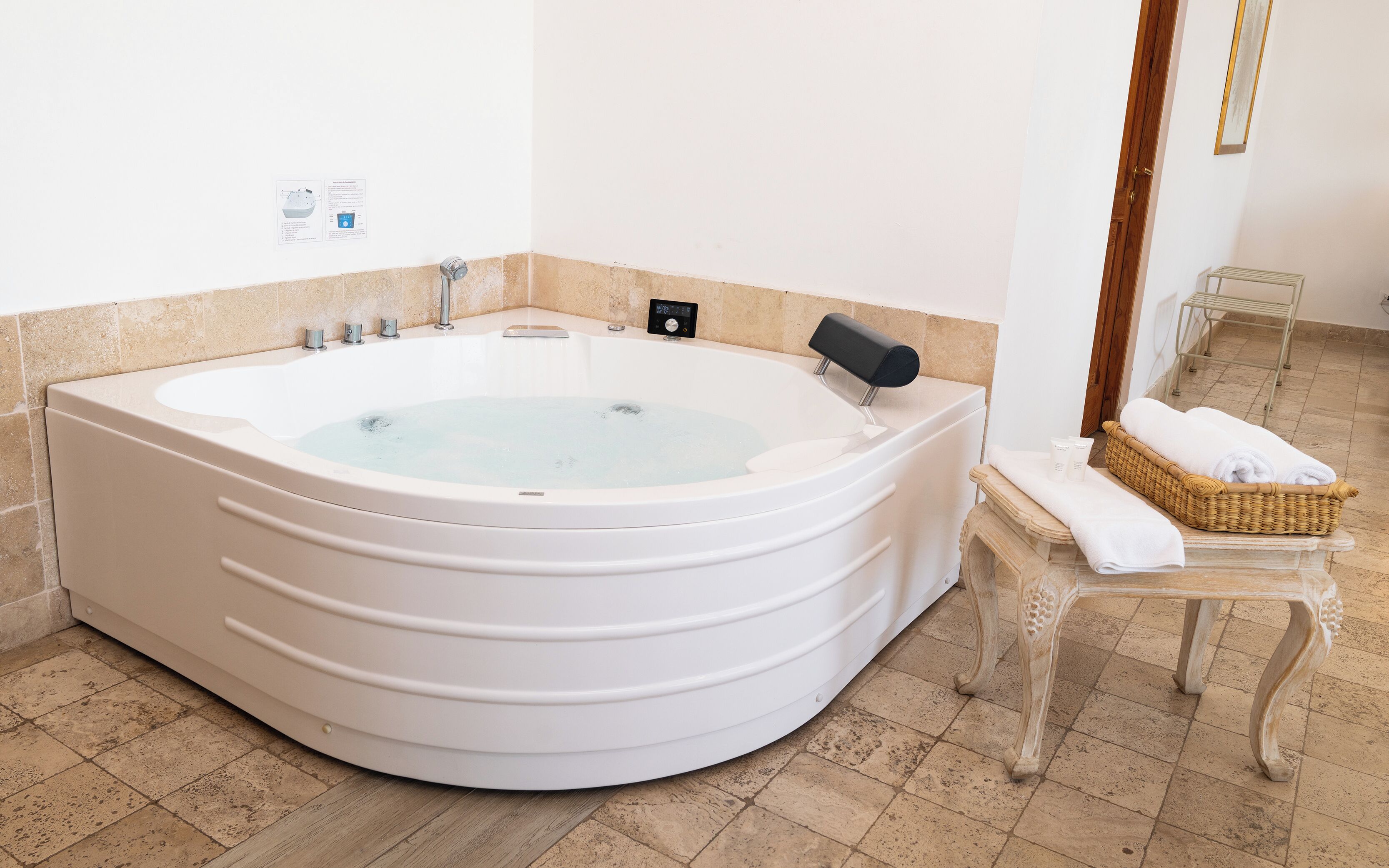 Una bañera blanca grande e independiente con un panel de control, una mesita con toallas y una cesta de mimbre.