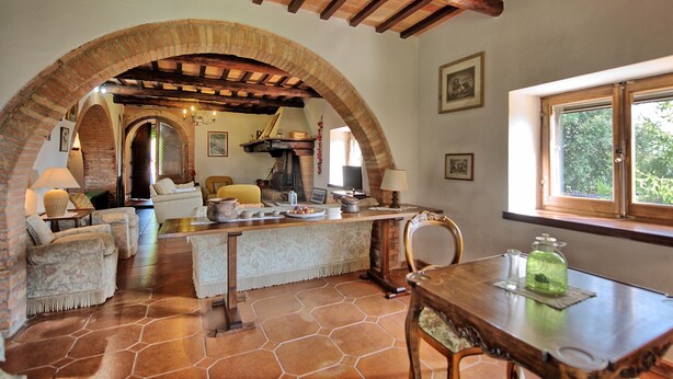 Un interior rústico con suelo de piedra, vigas de madera, una mesa de comedor con sillas y una zona de cocina con chimenea.