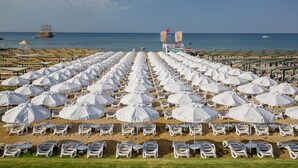A beach with rows of white sun loungers and umbrellas, a ship in the distance, and a clear sky. - Side Sunport Hotel & Spa - All Inclusive (Manavgat)