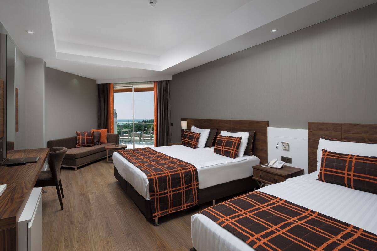 A hotel room with a large bed, a smaller sofa, a desk with a TV, and a balcony with a view.