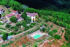 An aerial view of a rural area with a small cluster of buildings, including a house with a red door and a white building with a red roof. There is a green field and a fenced area with a pool.