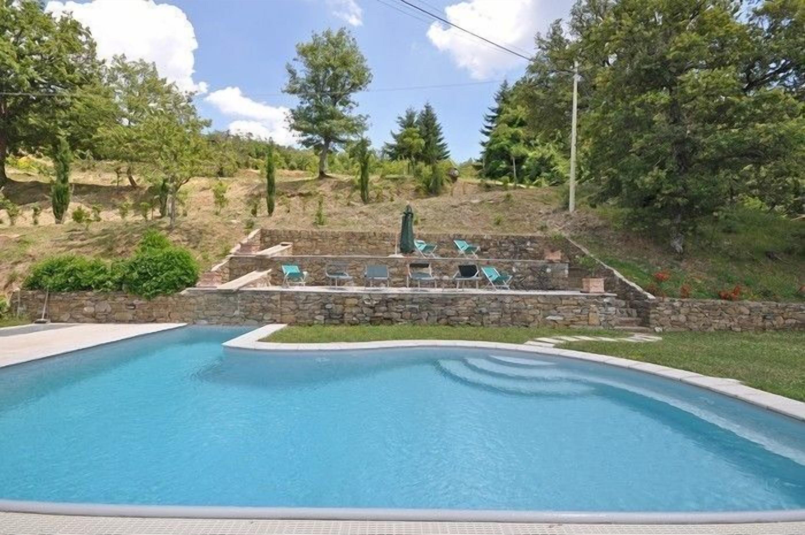 Ein Schwimmbecken mit Steinmauer und Liegestühlen, umgeben von Bäumen und einer Steintreppe.