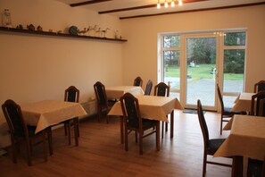 Un comedor con mesas y sillas de madera, un estante con objetos decorativos y vistas al exterior a través de un ventanal grande.