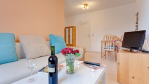 Ein gemütliches Wohnzimmer mit einem weißen Sofa, blauen Kissen, einem kleinen Tisch mit einer Weinflasche, einer Vase mit Blumen und einem Fernseher auf einem Holzboden.