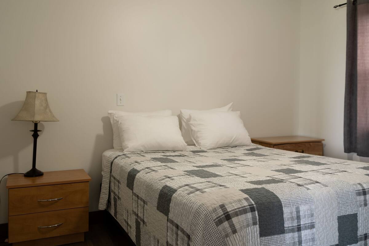 Un dormitorio con una cama, dos mesitas de noche, una lámpara y un ventanal con cortinas.