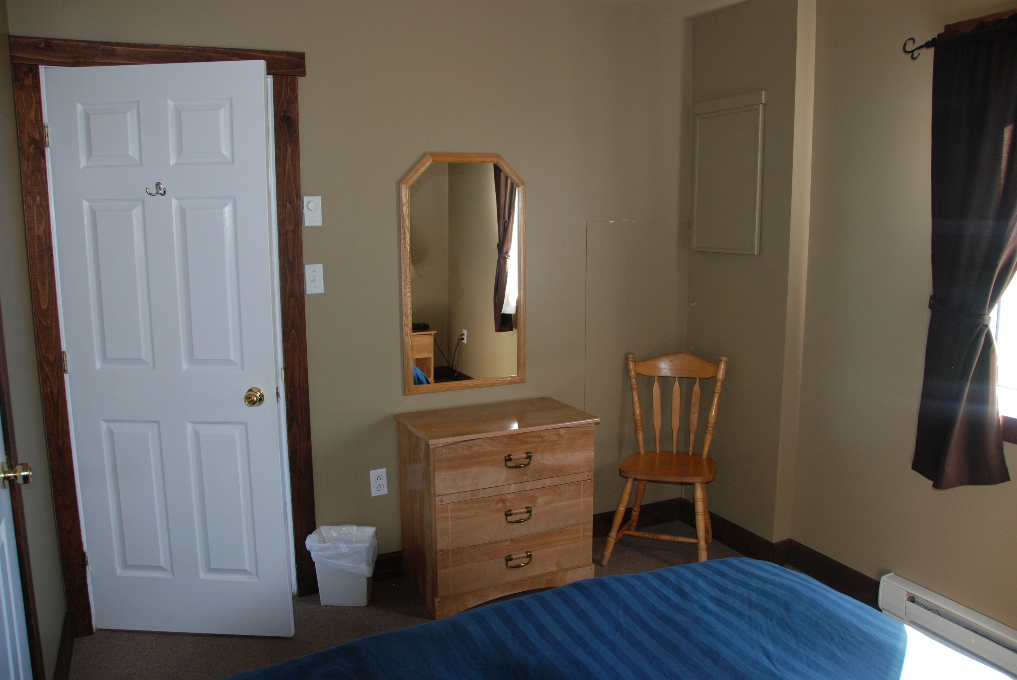 A bedroom with a bed, a wooden dresser, a chair, a mirror, and a door.