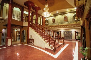 Una imponente escalera con barandillas ornamentales y un candelabro, que conduce a un espacio en el segundo piso con paredes decorativas y un espejo grande.
