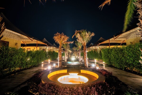 Vista nocturna de un complejo turístico con una fuente circular central, palmeras y senderos bien iluminados.