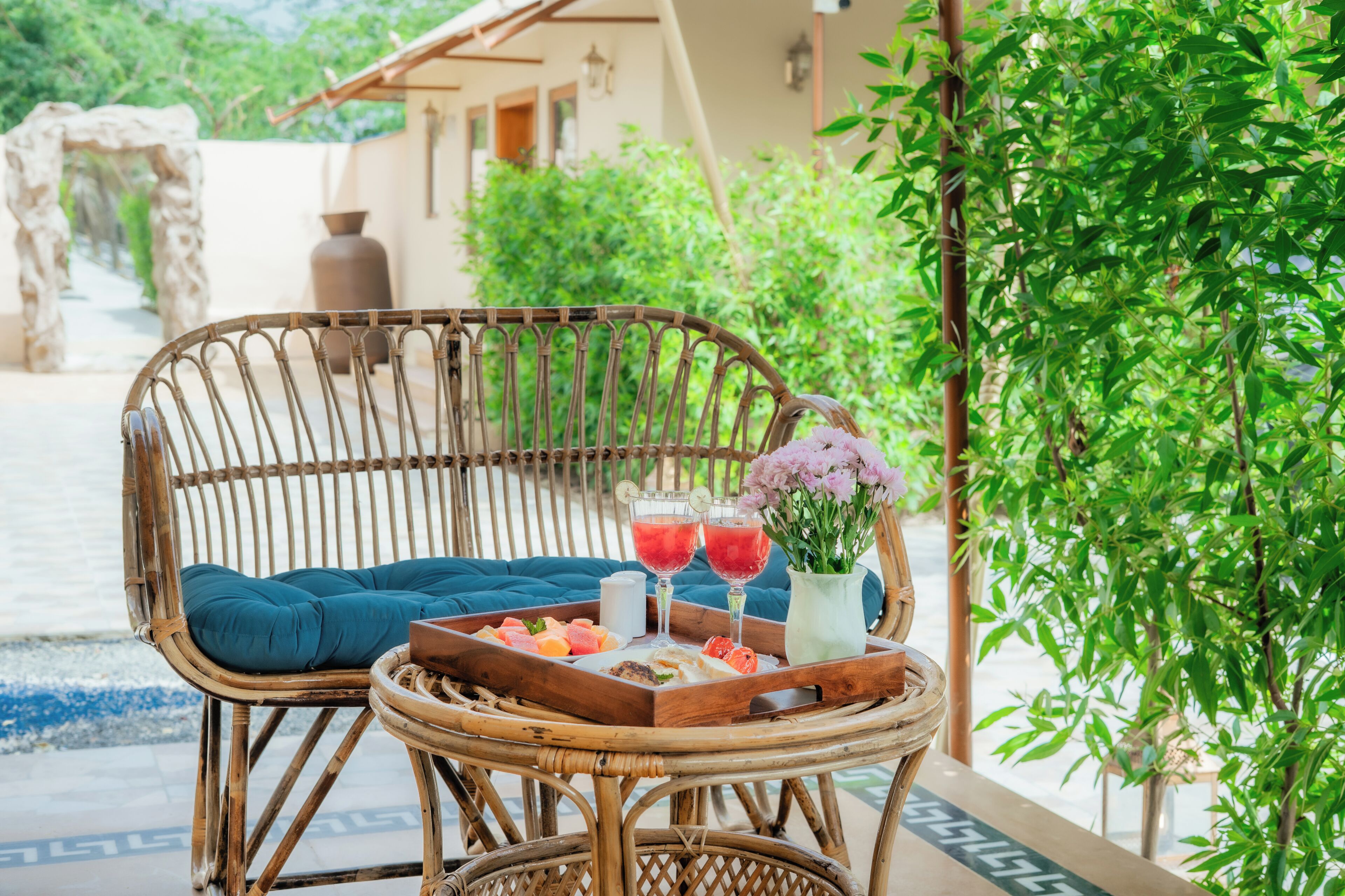 A wicker chair with a blue cushion, a tray with fruit and drinks, and a vase with flowers.