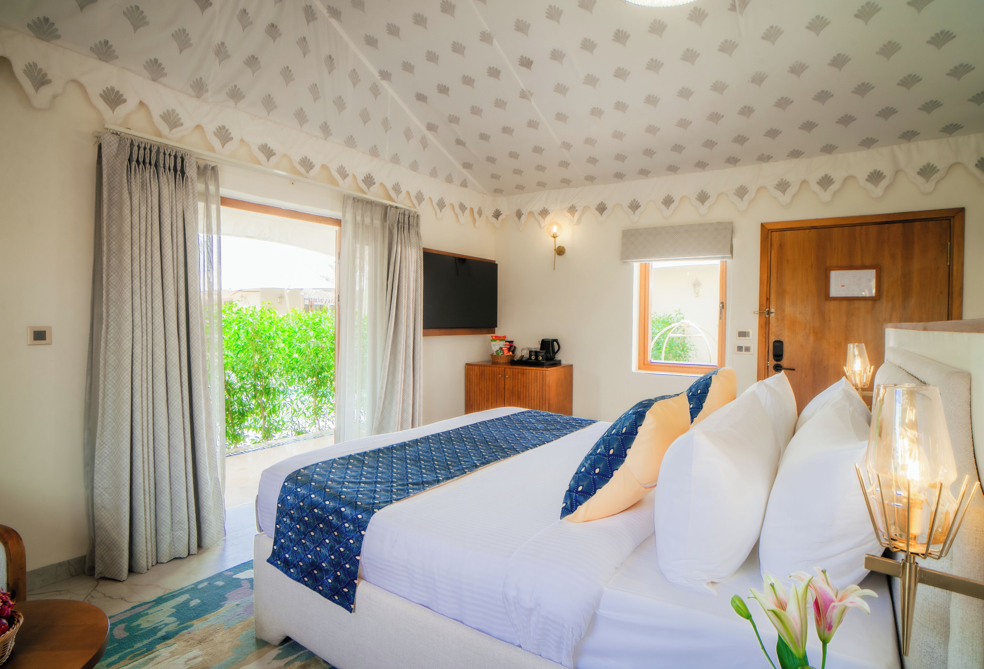 Habitación de hotel con cama, cortinas, televisor, puerta de madera y ventana con vista a follaje.