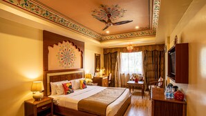 A hotel room with a large bed, a desk, a chair, a TV, and a ceiling fan. - Chokhi Dhani The Palace Hotel (Jaisalmer)