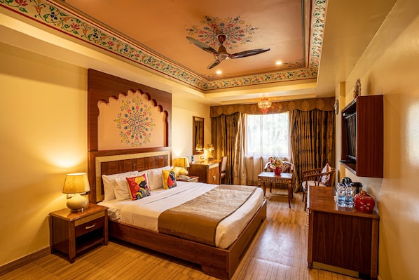 A hotel room with a large bed, a desk, a chair, a TV, and a ceiling fan. - Chokhi Dhani The Palace Hotel (Jaisalmer)