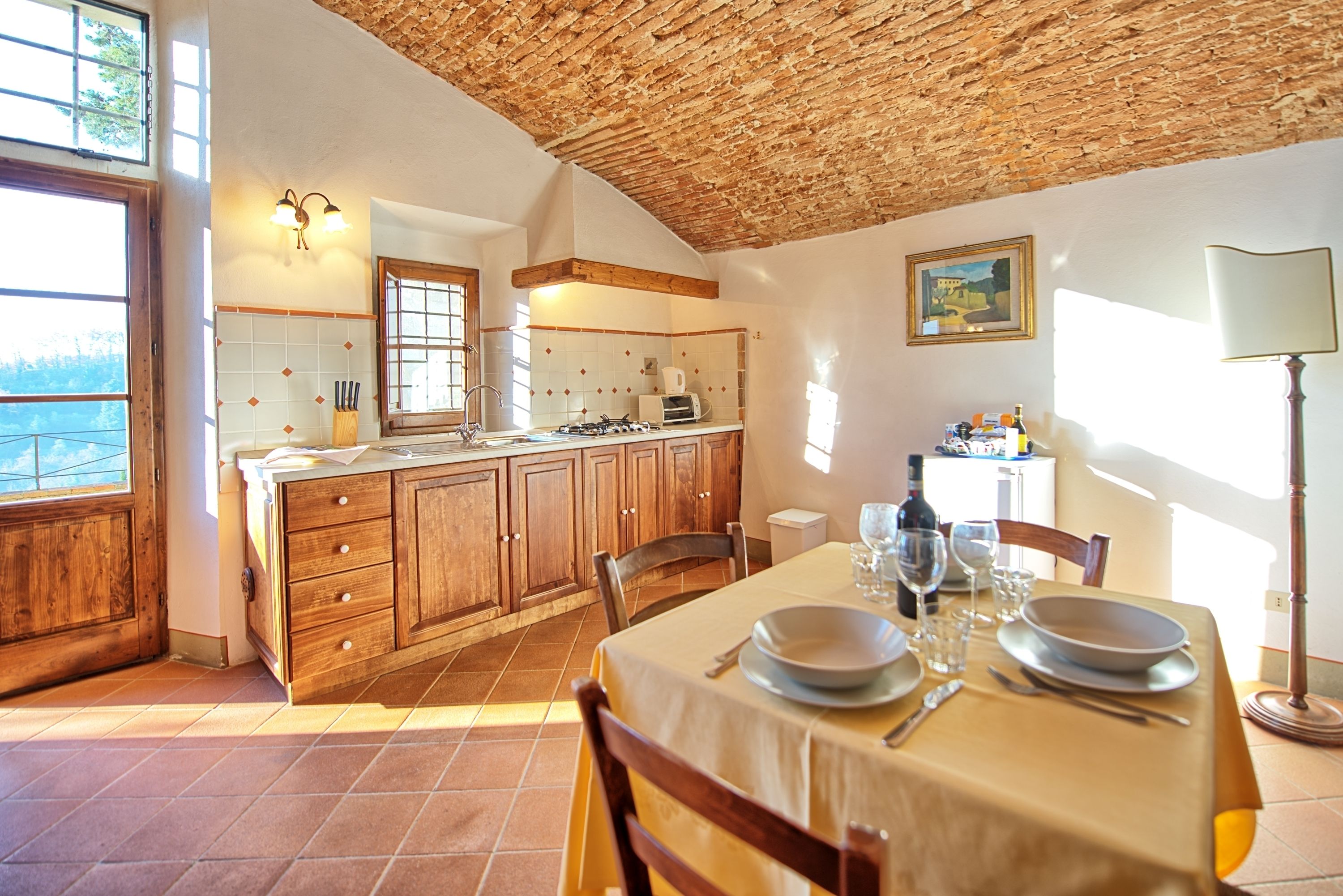 A dining area with a table set for two, wooden cabinets, a kitchen in the background, and a view through a window.