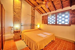 Un dormitorio con pared de ladrillo, vigas de madera en el techo, una cama con colcha amarilla, dos lámparas de noche y una ventana con contraventanas de madera.