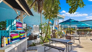 Un espacio al aire libre con patio, mesas y sillas de madera, un bar junto a la piscina con toallas de colores y un edificio azul al fondo.