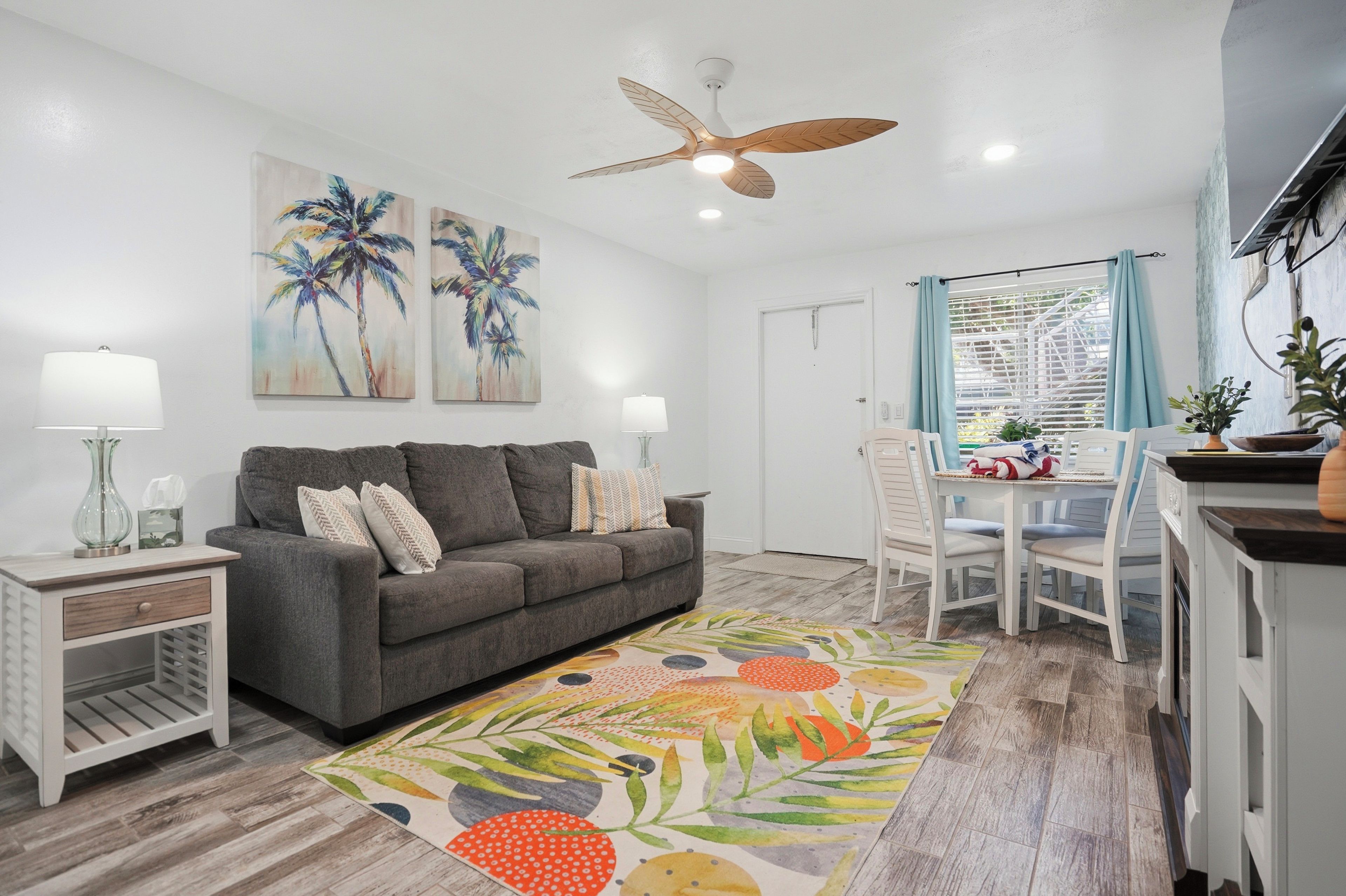 A modern living room with a grey sofa, two wall-mounted lamps, a ceiling fan, and a dining area with a white table and chairs.