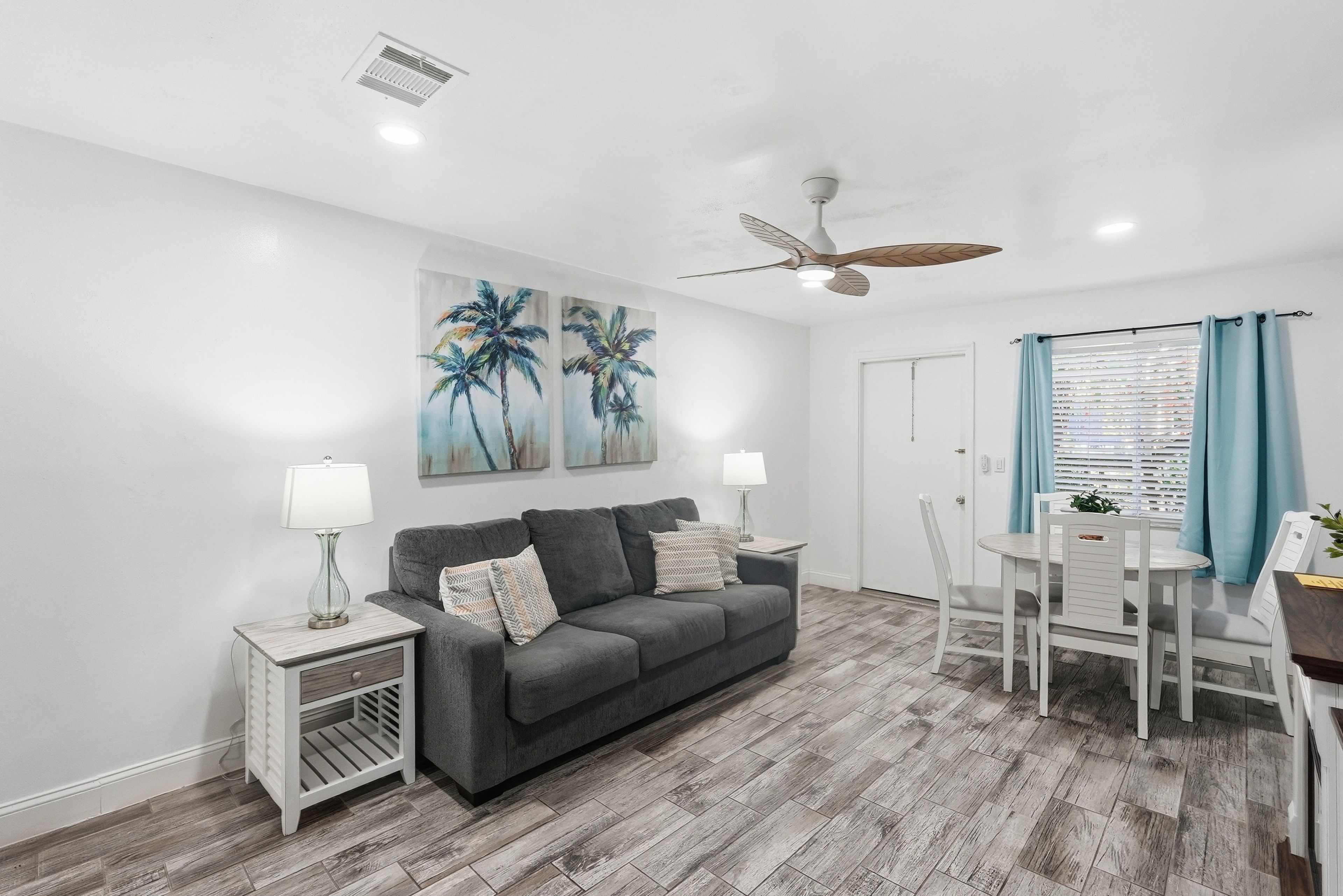 A modern living room with a grey sofa, two side tables with lamps, a ceiling fan, and artwork of palm trees.