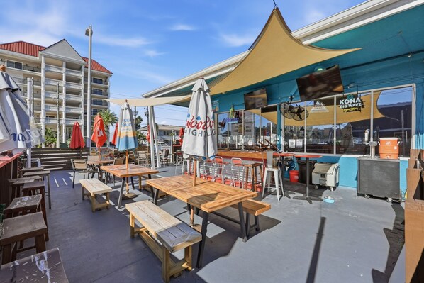 An outdoor seating area with wooden tables and benches, umbrellas, and a bar with a blue awning. - Malibu Resort Motel (North Redington Beach)