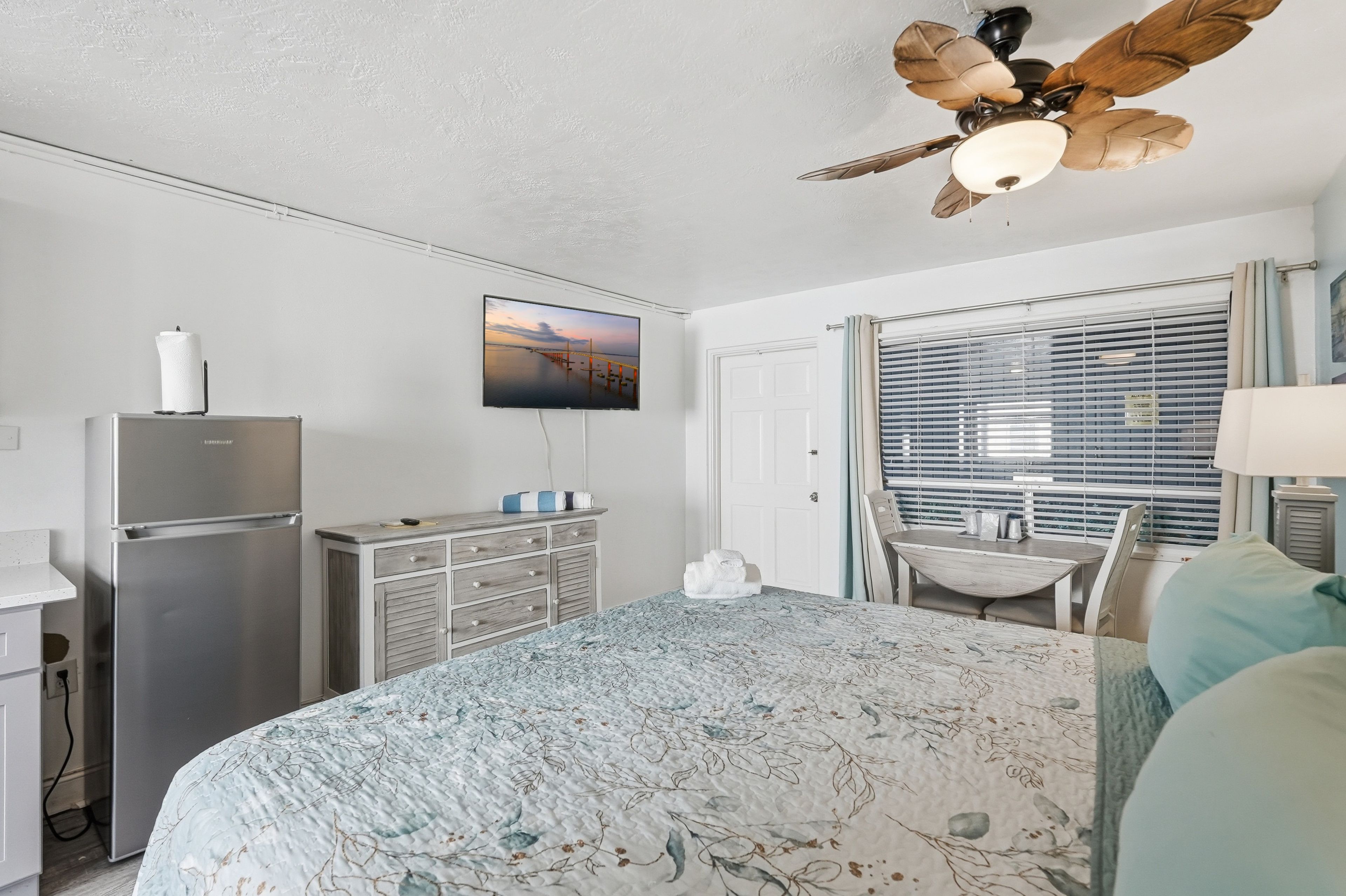 A bedroom with a bed, a television mounted on the wall, a ceiling fan, a window with blinds, and a refrigerator.
