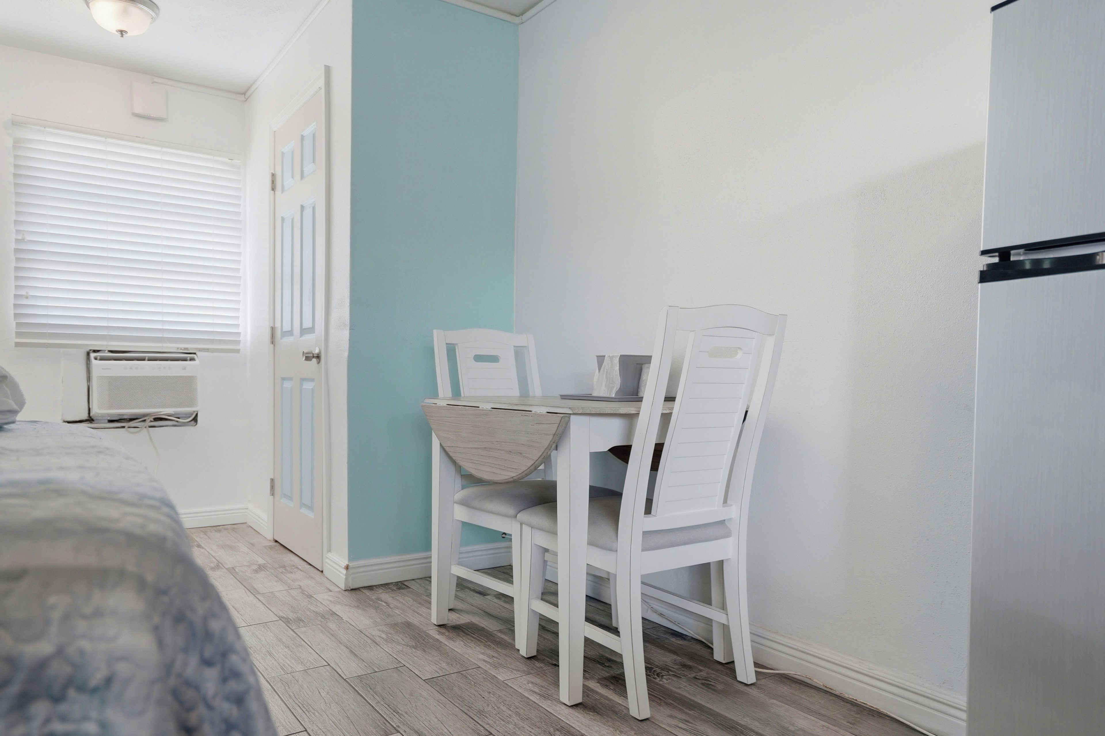 A small room with a bed, a table with chairs, a window with blinds, and a door.