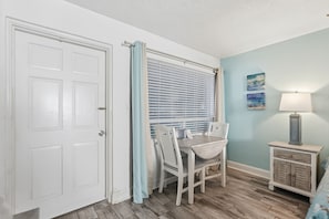 A room with a closed white door, a window with blinds, a table with chairs, a lamp, and a cabinet. - Malibu Resort Motel (North Redington Beach)