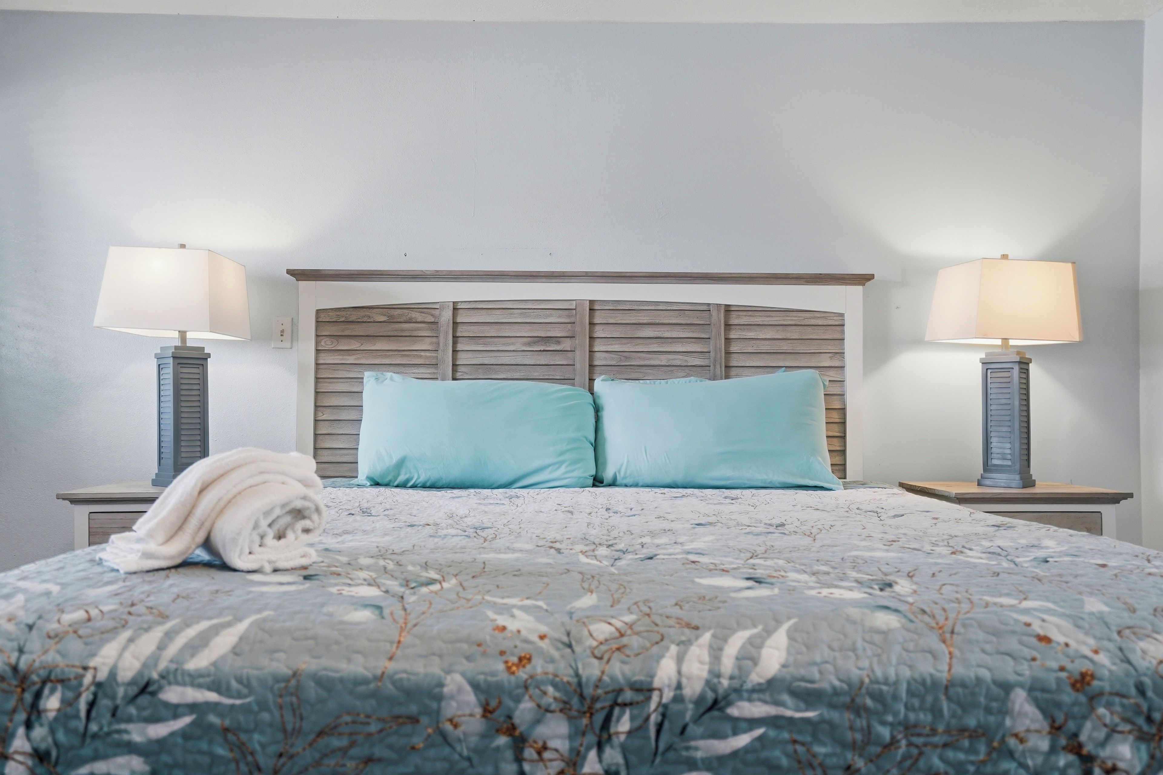Un dormitorio con cabecera de madera, dos lámparas de noche, una toalla enrollada y una colcha de estampado floral.