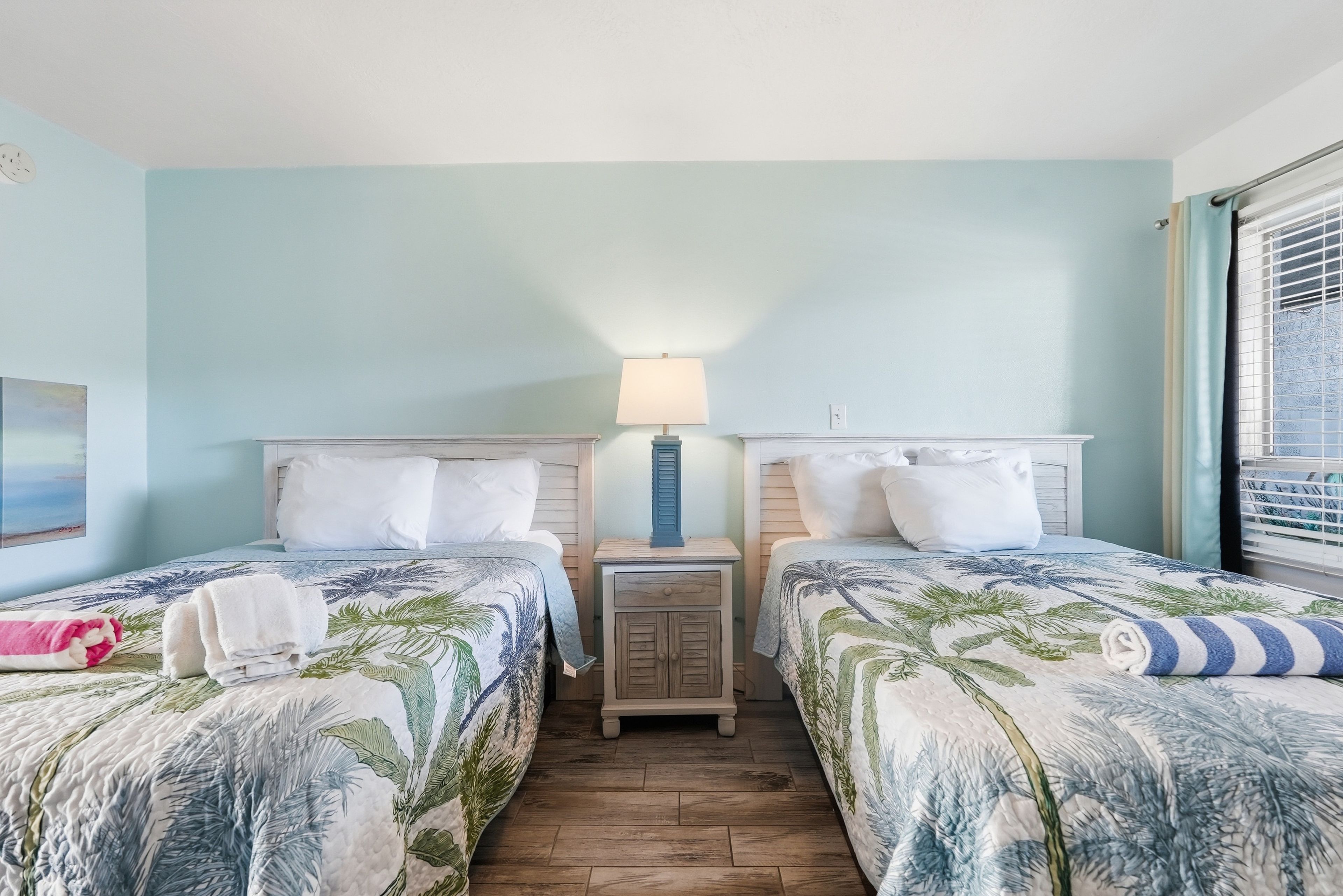 A room with two beds, each with a floral bedspread, a nightstand with a lamp, and a window with blinds.
