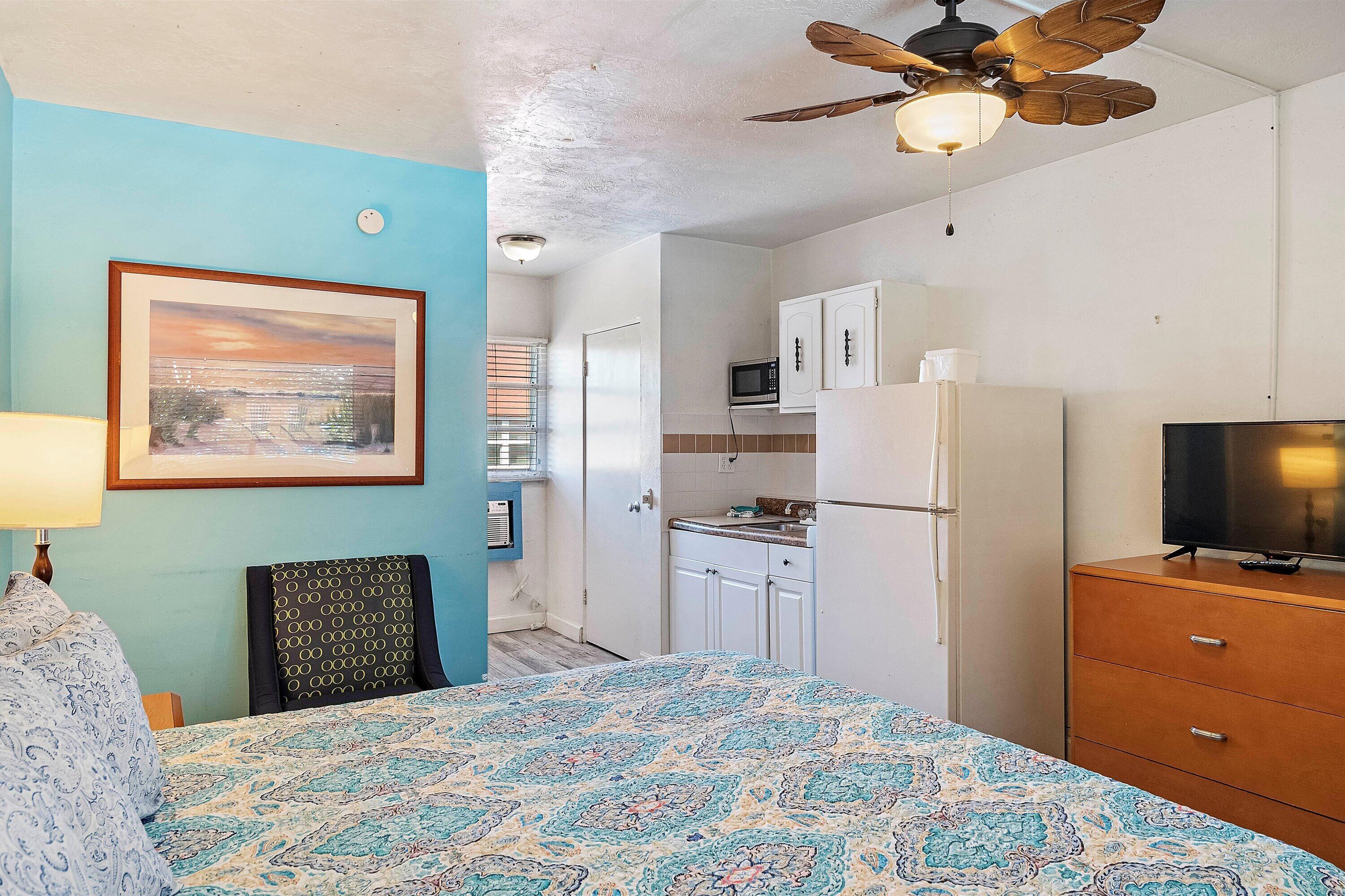 A bedroom with a bed, a ceiling fan, a TV, and a kitchenette.