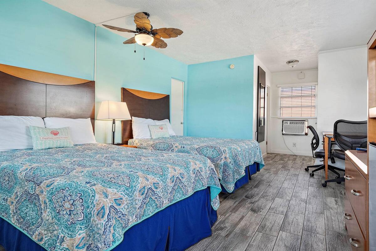 A hotel room with two beds, a desk, an air conditioning unit, and a ceiling fan.