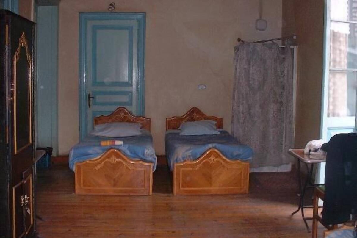 A room with two wooden beds, a blue door, a small table, and a window.