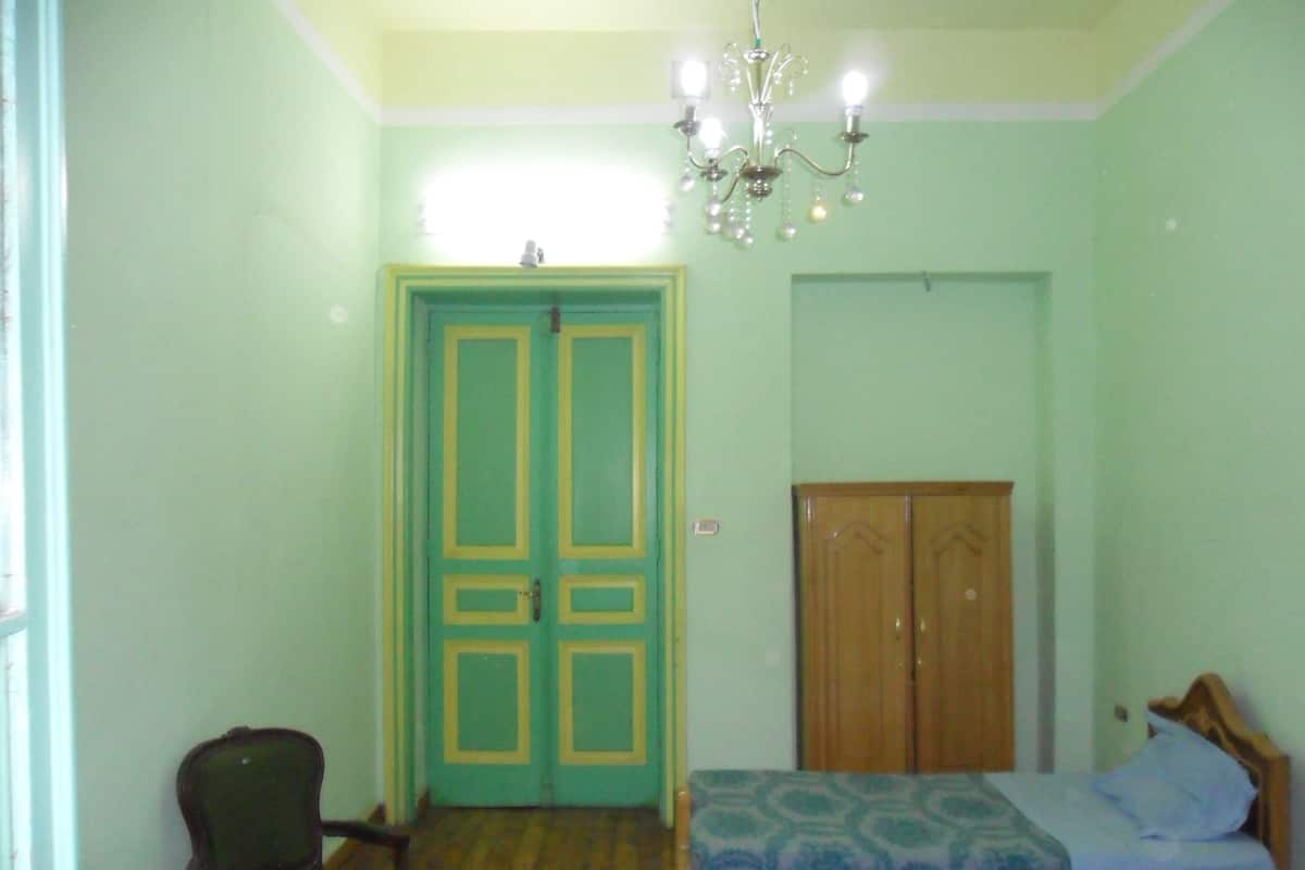 A room with a bed, a chair, a wooden wardrobe, and a chandelier.