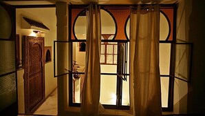 A room with a view through a window with curtains, a wooden door, and a small table with a plant. - Riad La Cigale (Marrakech)