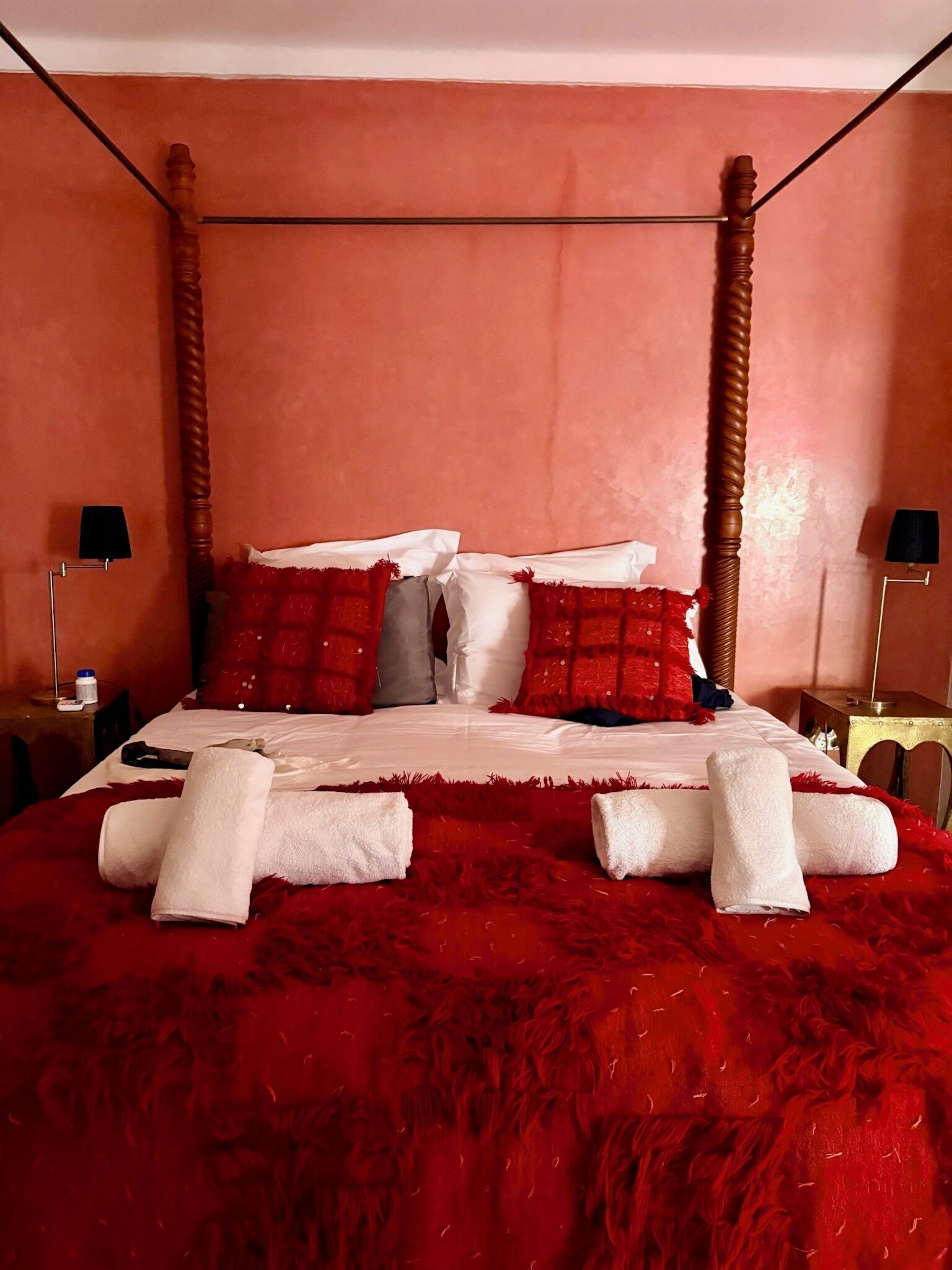 A four-poster bed with white linens and red pillows, a red furry blanket, and two bedside tables with lamps.
