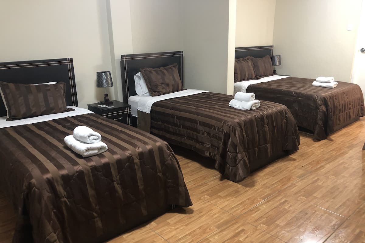 A hotel room with three beds, each with a brown bedspread and white pillows. There is a nightstand with a lamp and a small table between the beds.