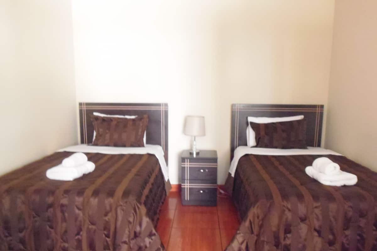 A hotel room with two beds, each with a pillow and a rolled towel. There is a nightstand with a lamp between the beds.