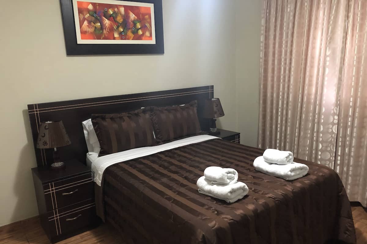 A neatly made bed with a brown striped bedspread, two white towels, a nightstand with a lamp, and a framed abstract painting on the wall.